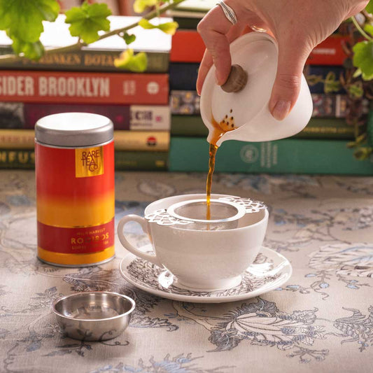 A person pouring rooibos tea from a gaiwan into a teacup and saucer, through a strainer. There is a tin of Rare Tea Company South African Wild Rooibos tea and a dish for the strainer to sit in.