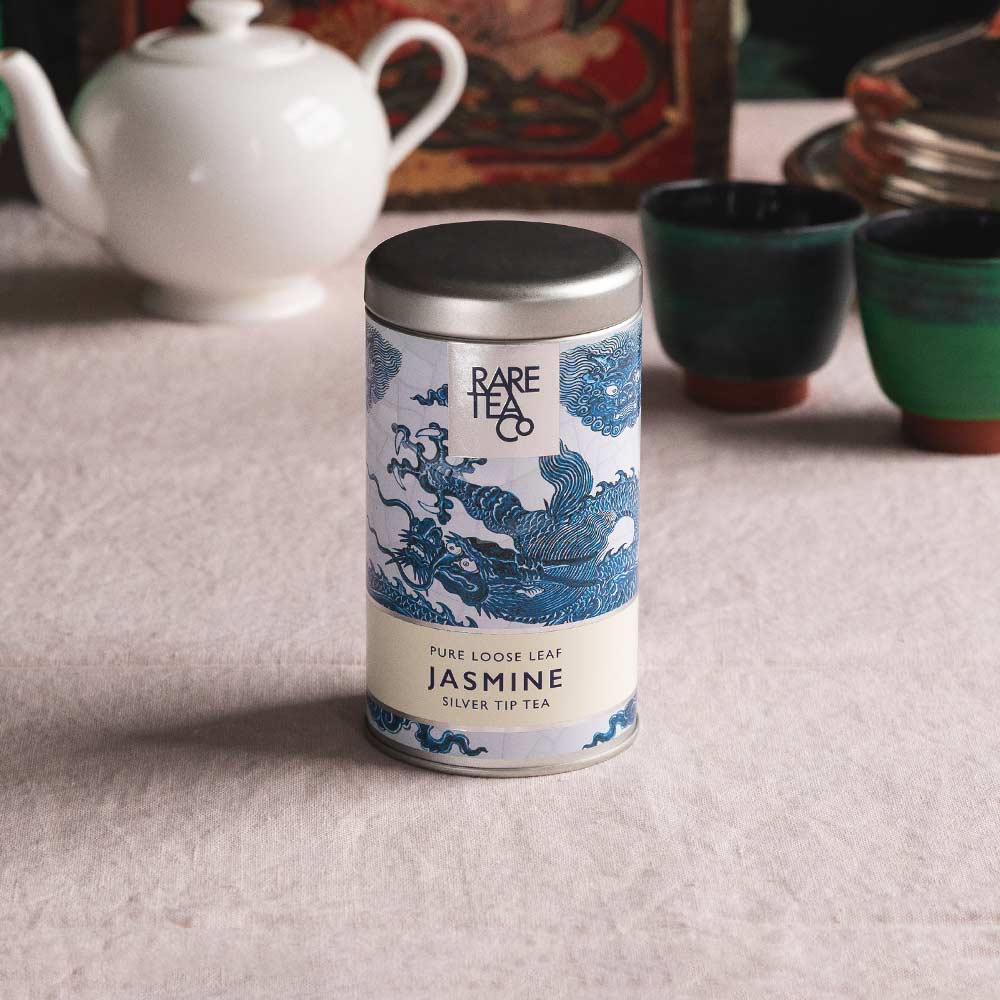 A tin of Rare Tea Company Silver Tip Jasmine tea on a white tablecloth with vintage teaware in the background.
