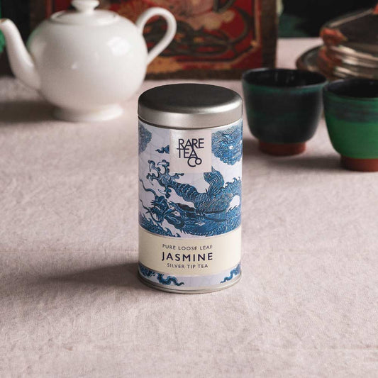 A tin of Rare Tea Company Silver Tip Jasmine tea on a white tablecloth with vintage teaware in the background.