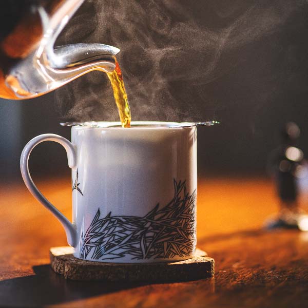 Steaming hot tea being poured into a white Rare Tea Company mug with black leaf patterns on a wooden surface.