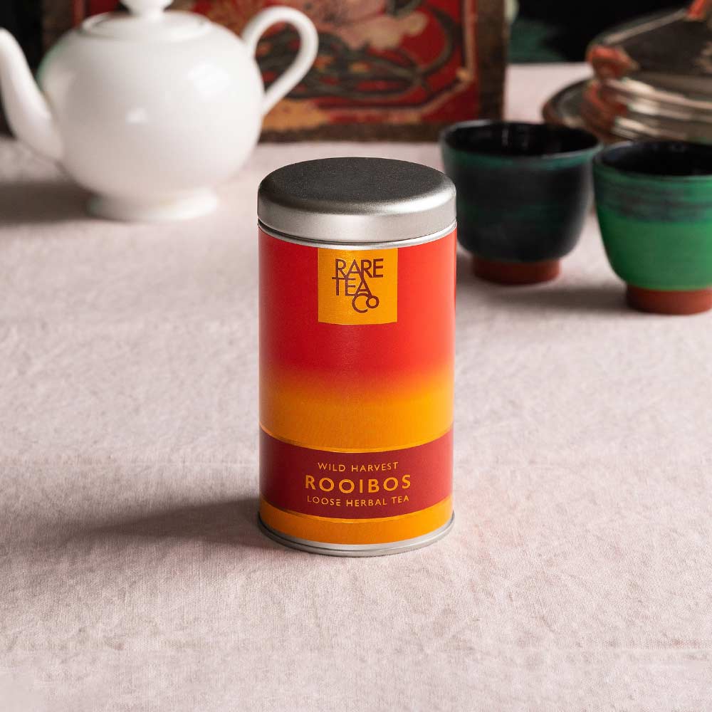 A tin of Rare Tea Company South African Wild Rooibos tea on a white tablecloth with vintage teaware in the background.